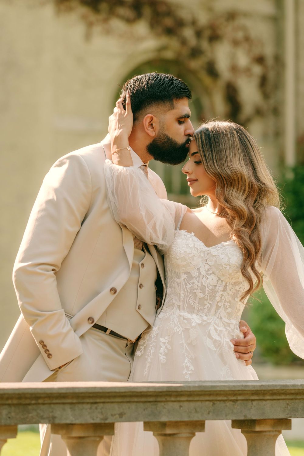 A romantic wedding couple embraces on a stone balcony, with the bride in white lace and groom in beige suit.