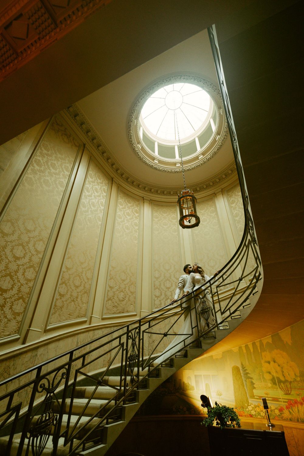 Elegant spiral staircase with ornate iron railing leads to domed skylight in a historic interior with patterned wallpaper.