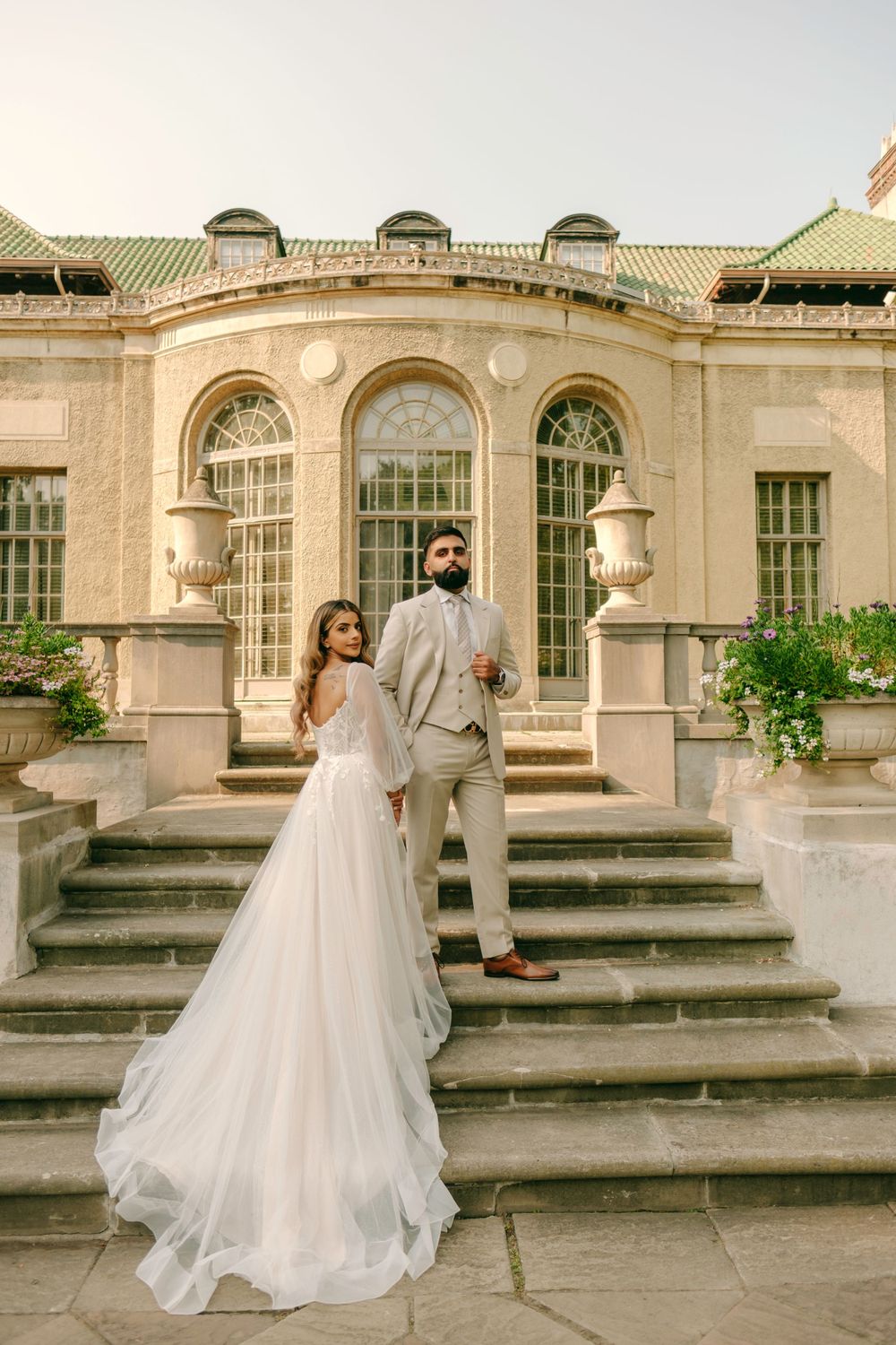 A wedding couple stands on stone steps in front of an elegant historic building with arched windows and columns.