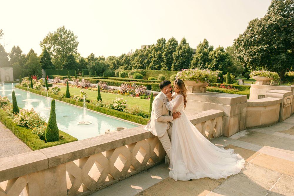 Romantic couple embraces on stone terrace overlooking manicured garden with fountains at sunset.