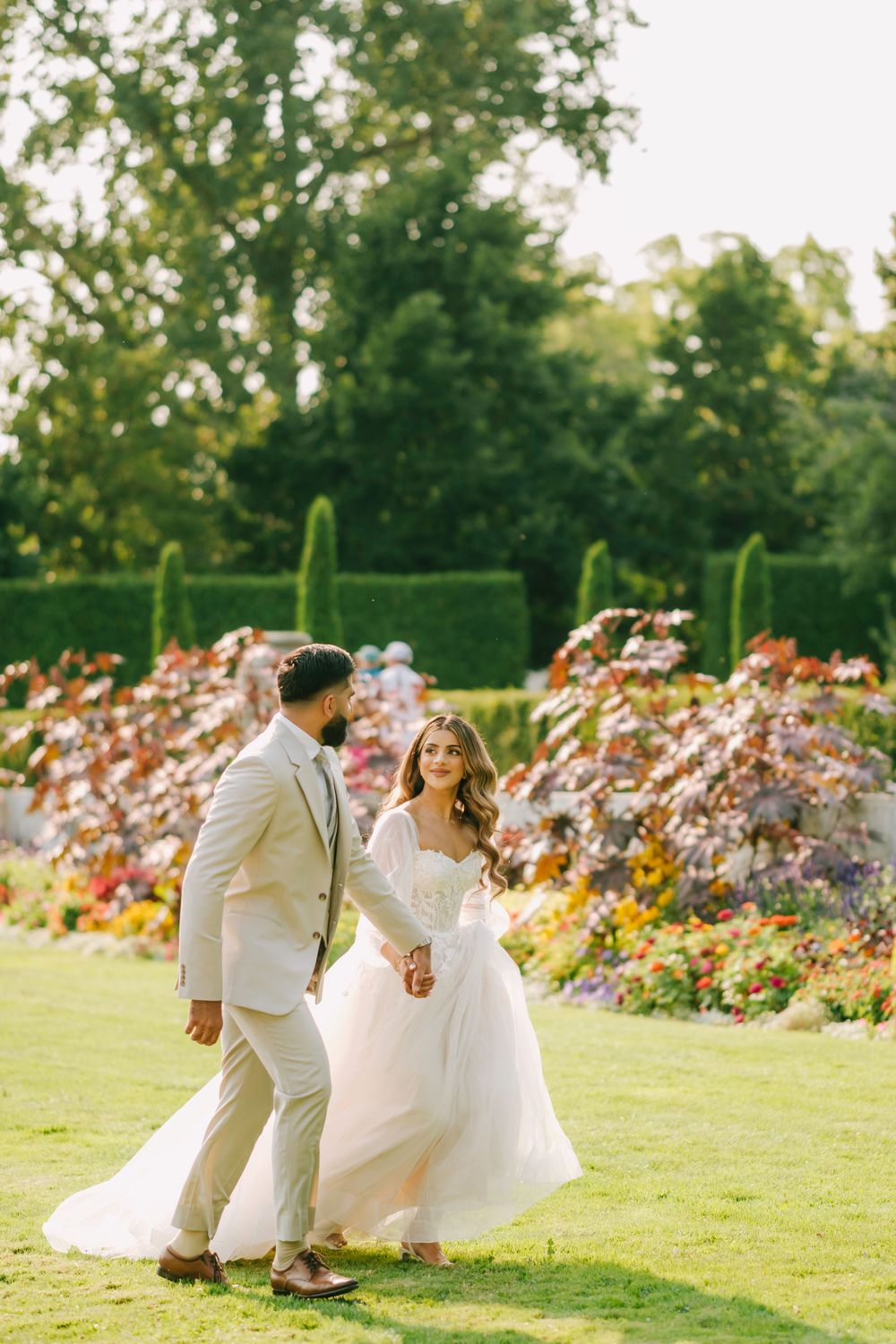 A couple in wedding attire stroll through a lush garden with vibrant flower beds and manicured hedges on a sunny day.