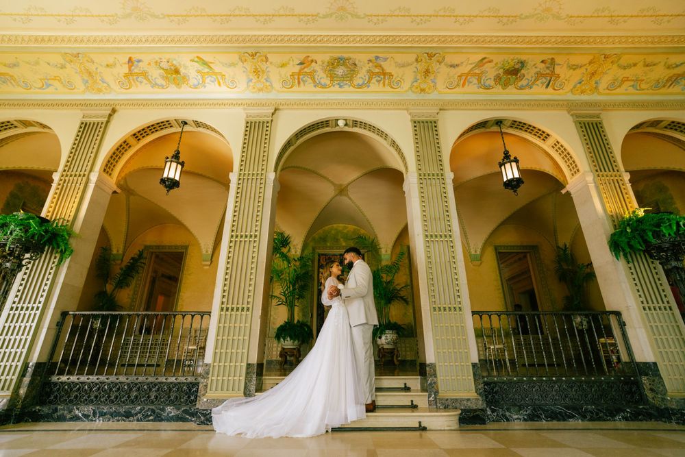Elegant wedding couple embraces on grand ornate veranda with arched columns and wrought iron railings.