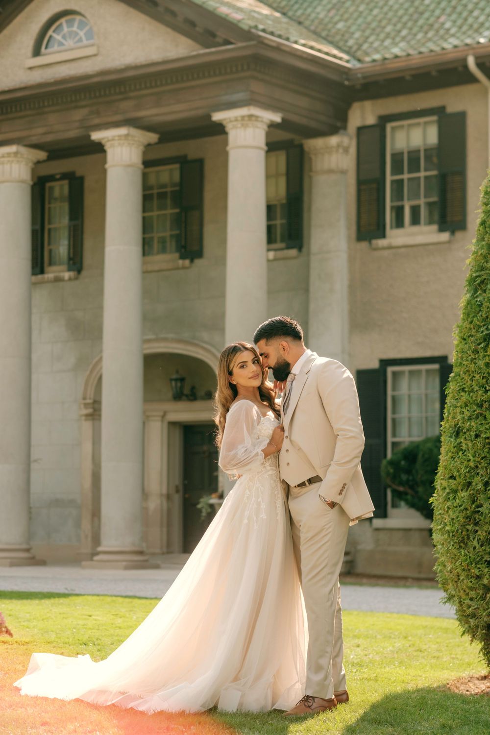 A romantic wedding couple embraces on the lawn of an elegant mansion with white columns and green-tiled roof.