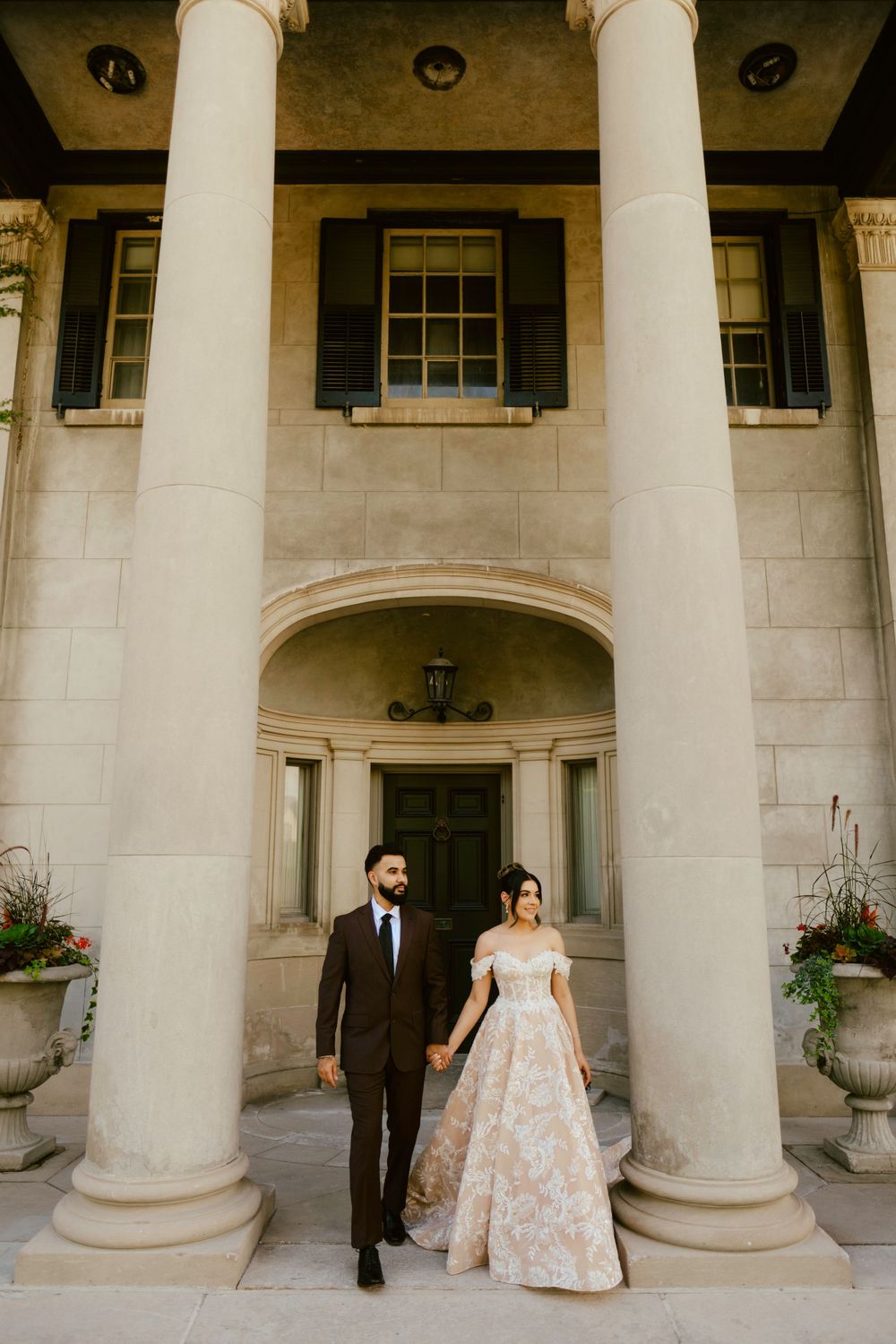 A couple stands between grand white columns at a stately mansion entrance wearing formal wedding attire.