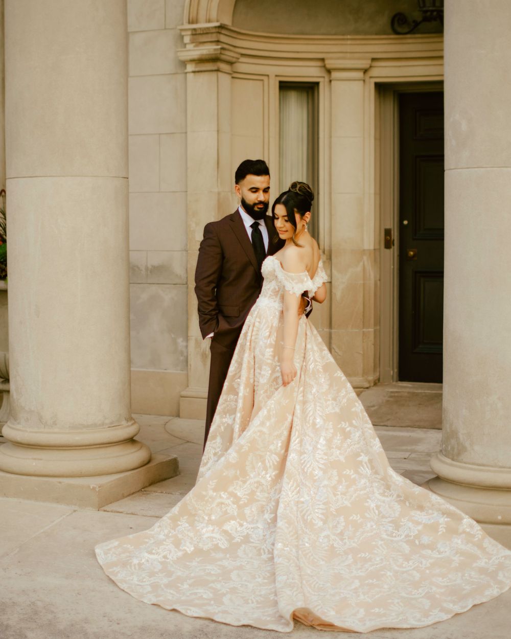 An elegant couple poses by white mansion columns showcasing a flowing cream lace wedding gown with long train.