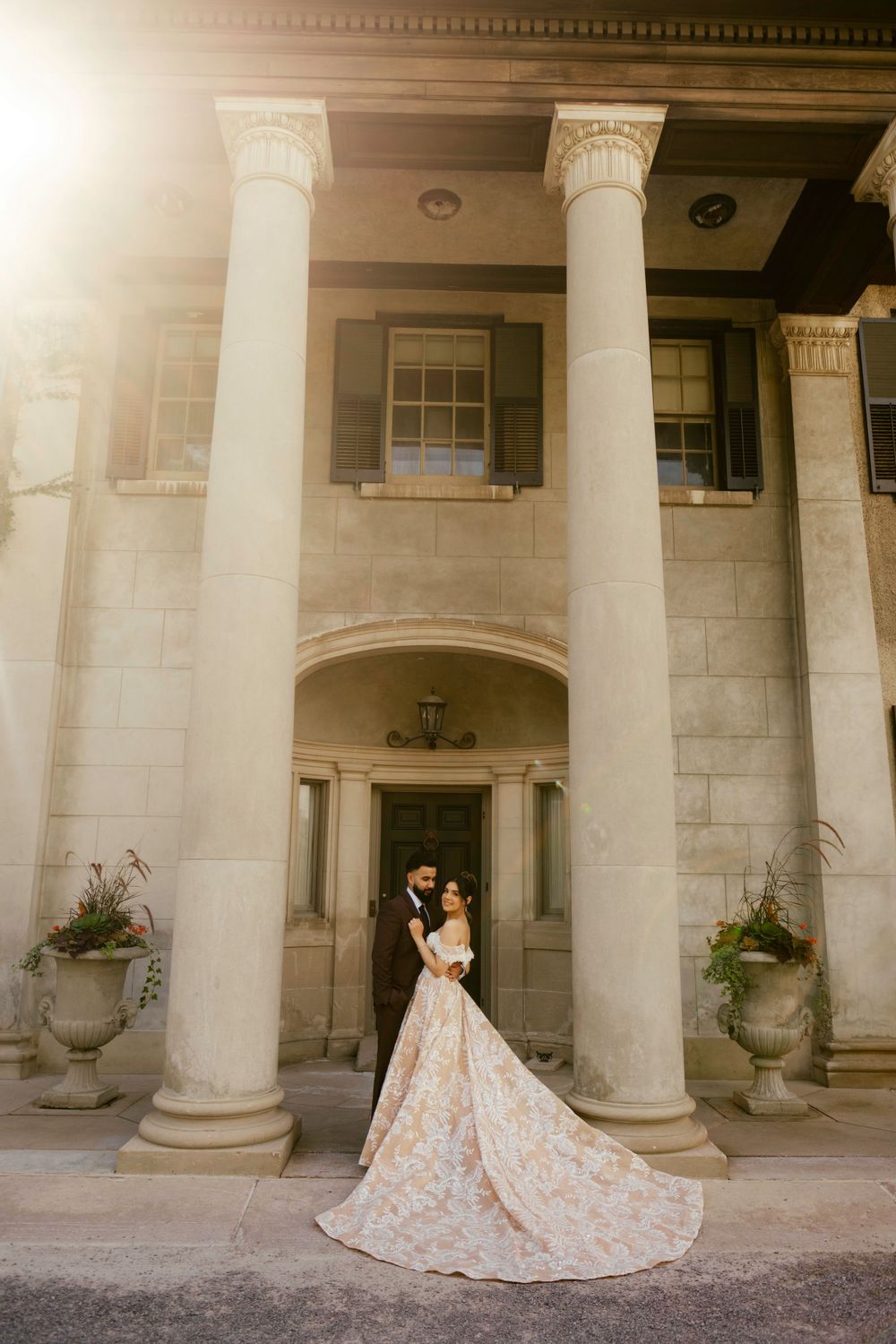 A couple poses between elegant white columns at a neoclassical mansion entrance with a flowing floral wedding gown.
