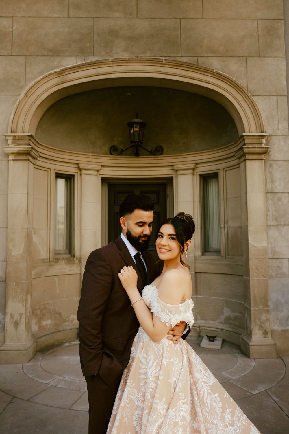 A romantic wedding photo in an elegant stone archway featuring a couple in dark suit and beige lace gown.
