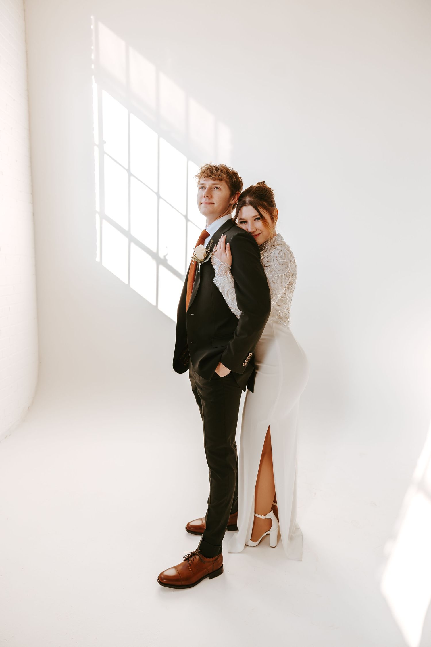 A couple poses elegantly in wedding attire against a bright white backdrop with dramatic window lighting.