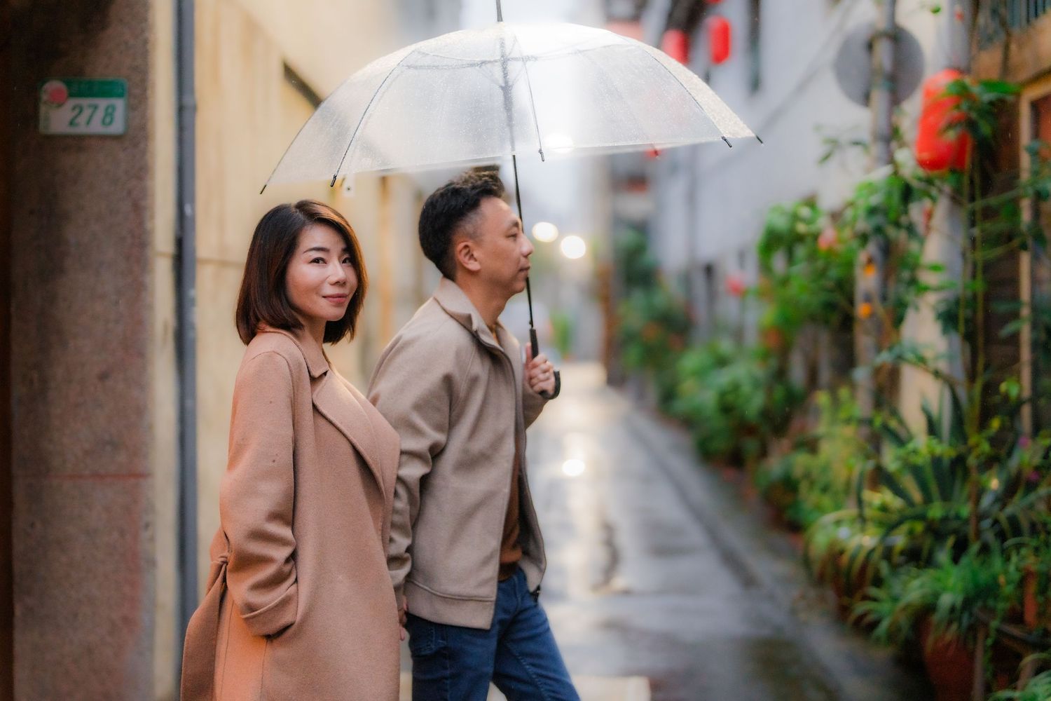 Two people walking with a white umbrella down a narrow alley on a rainy day in Japan while wearing beige coats.