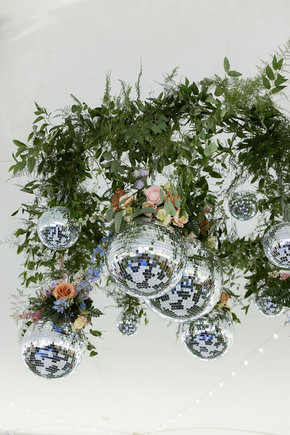 Elegant wedding ceiling decor featuring lush greenery, pink flowers, and hanging mirrored disco balls against a white backdrop.