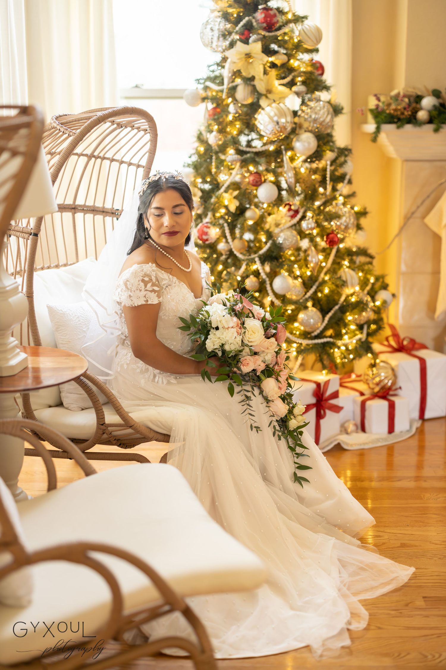 A bride in lace dress poses on rattan chair with bouquet near decorated Christmas tree and wrapped presents.