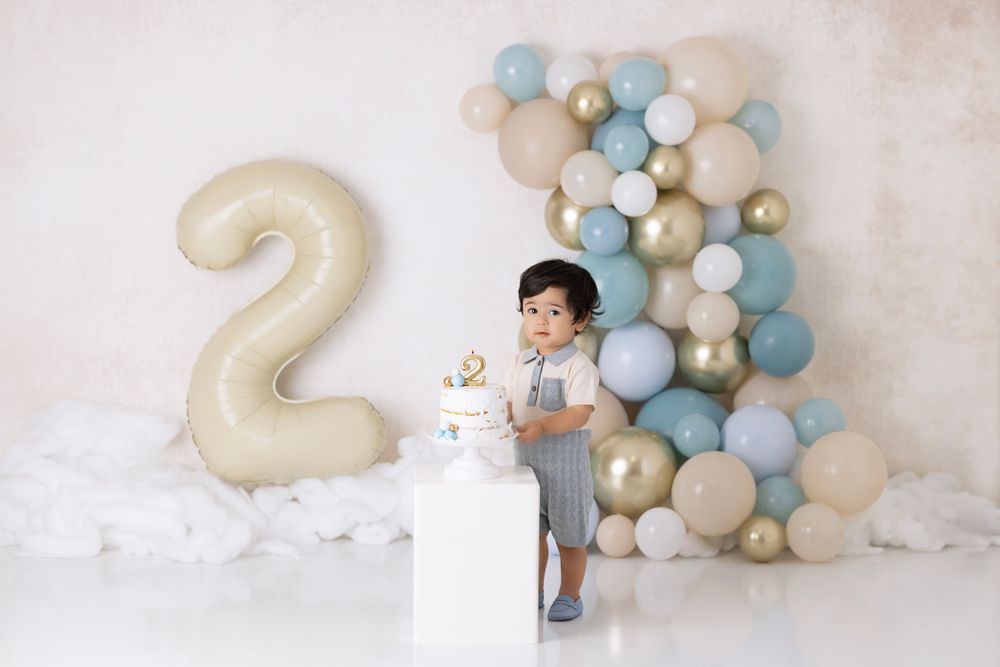 Birthday celebration setup with large number 2 balloon, blue and gold balloon arch, and white decorative pedestal against white backdrop.