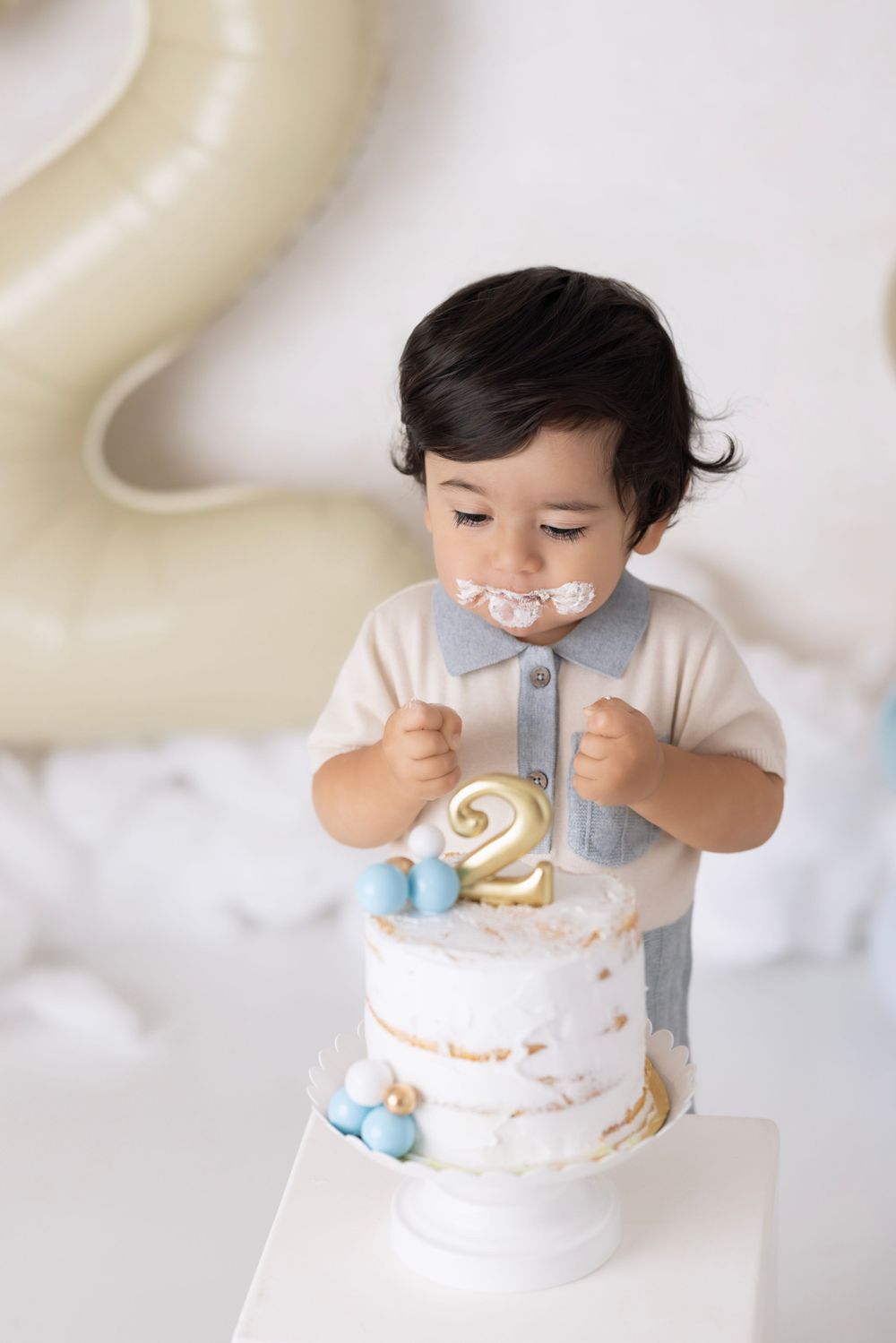 A toddler stands in front of a white birthday cake decorated with a gold number 2 and blue accents.