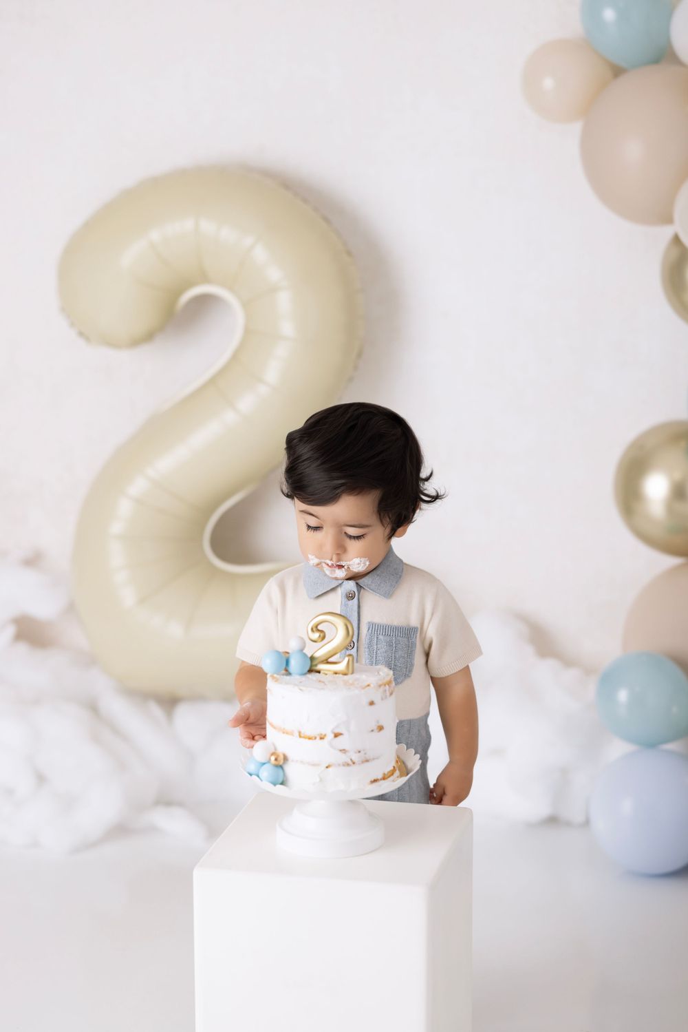 Birthday setup with cream-colored number 2 balloon, white cake on pedestal, and pastel balloon arch decorations.