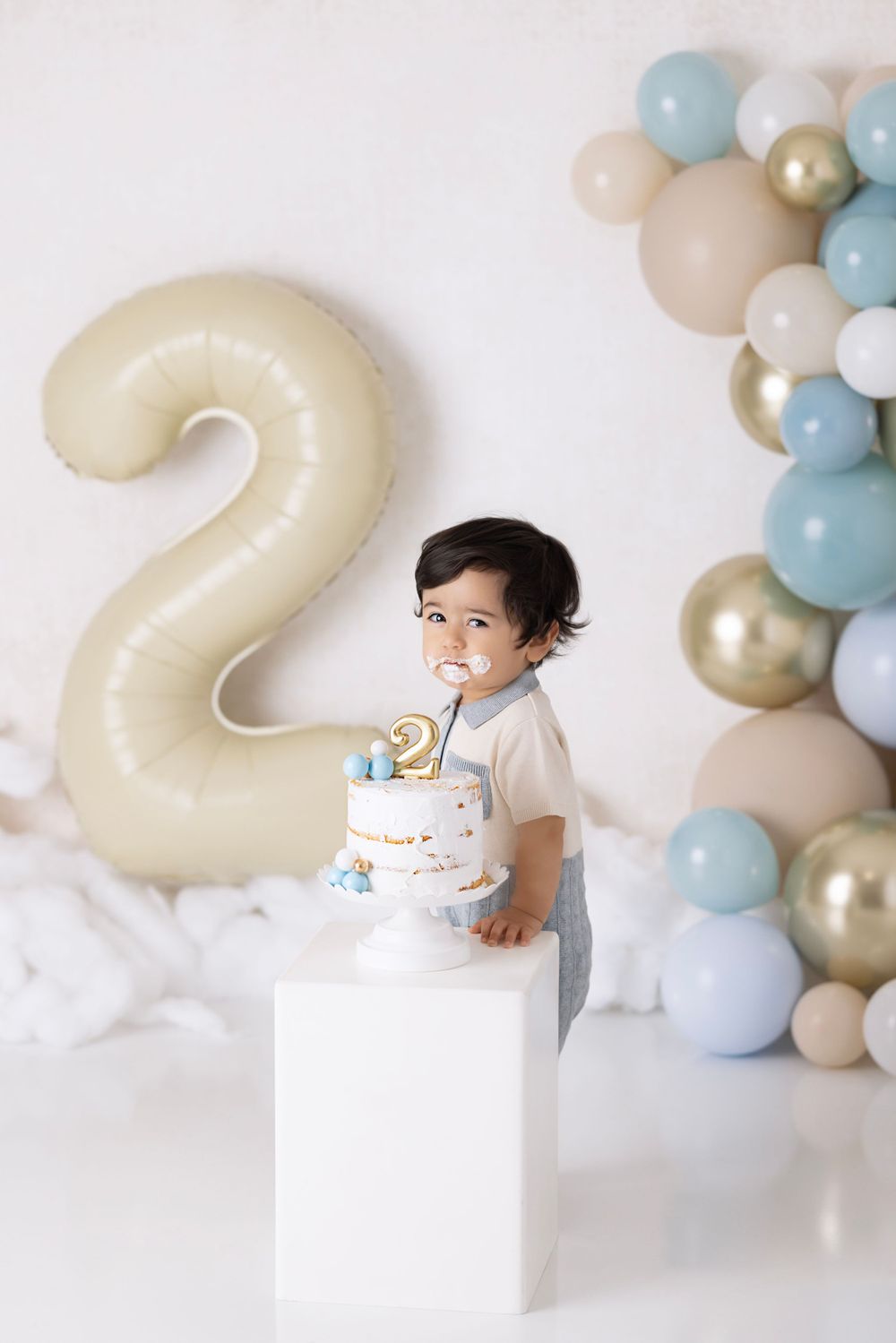 Second birthday party setup with white decorative cake, large cream-colored number 2 balloon, and pastel balloon arch.