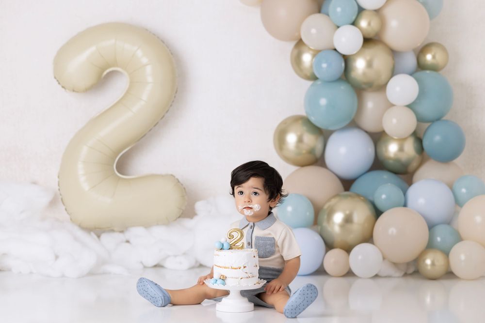 Birthday setup with large number 2 balloon, pastel blue and gold balloon arch, and white cake against white backdrop.