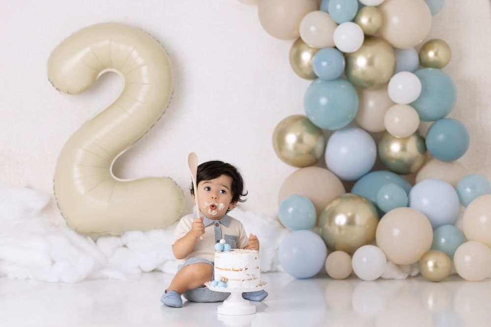Second birthday celebration scene with large '2' balloon, pastel balloon garland, and cake on white background.