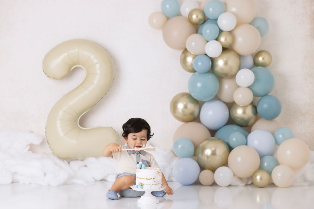 Birthday celebration setup with a large number 2 balloon and blue, white and gold balloon arch arrangement against white backdrop.