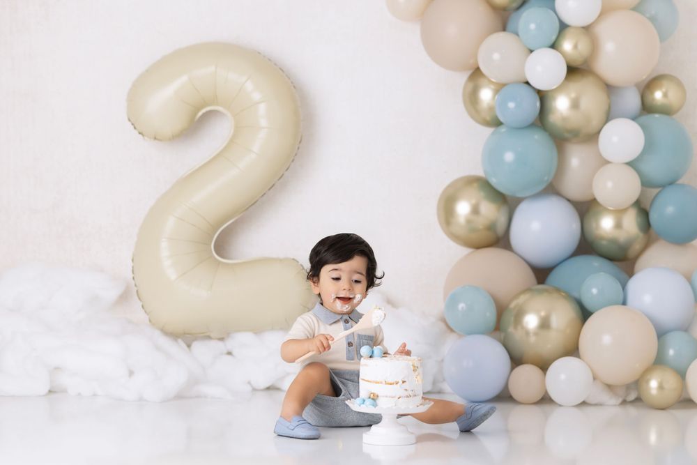 Birthday setup with large number 2 balloon, blue and gold balloon arch arrangement with cake and decorative elements on floor.