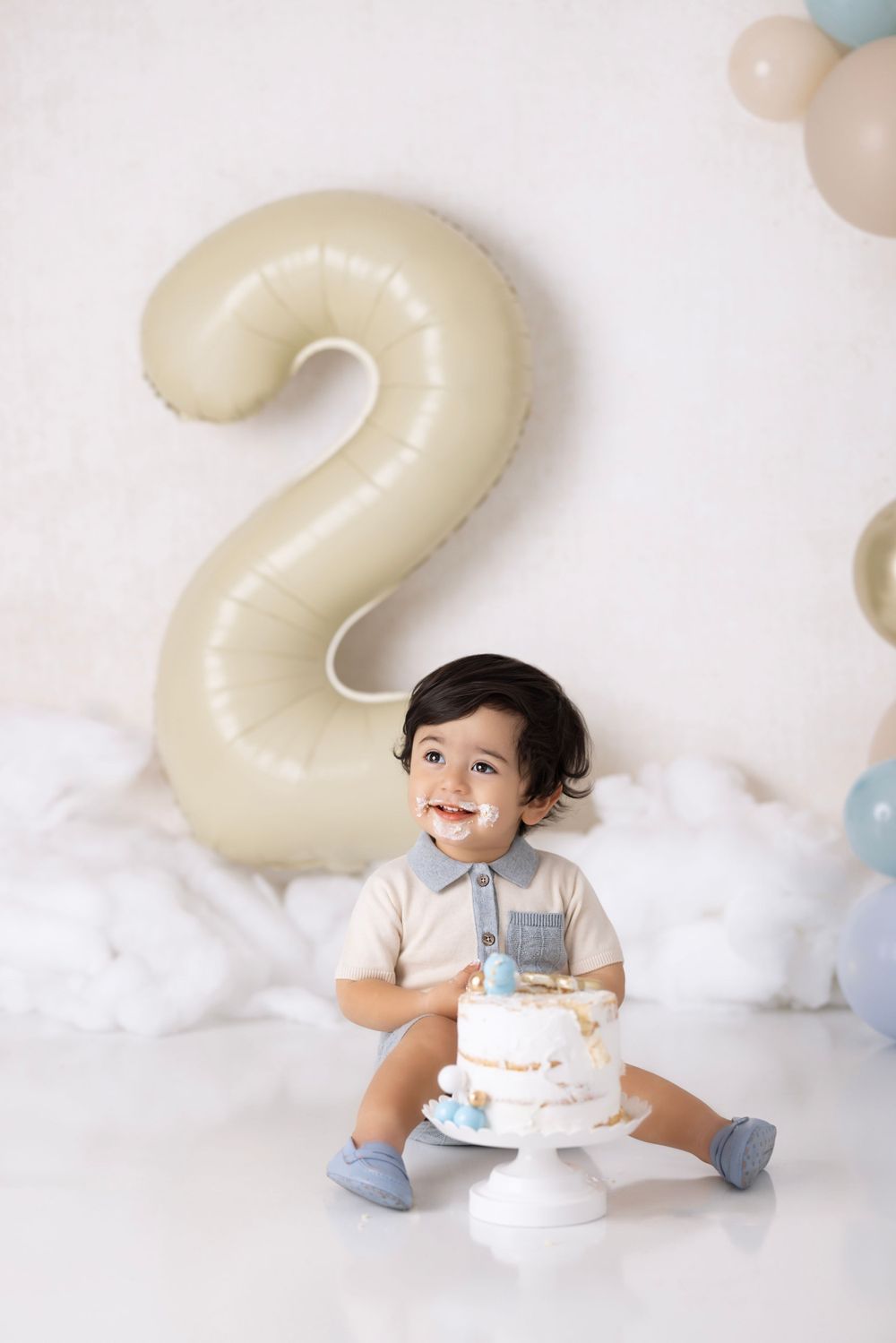 A toddler celebrates their second birthday with a cake and large number 2 balloon against a white cloudy backdrop.