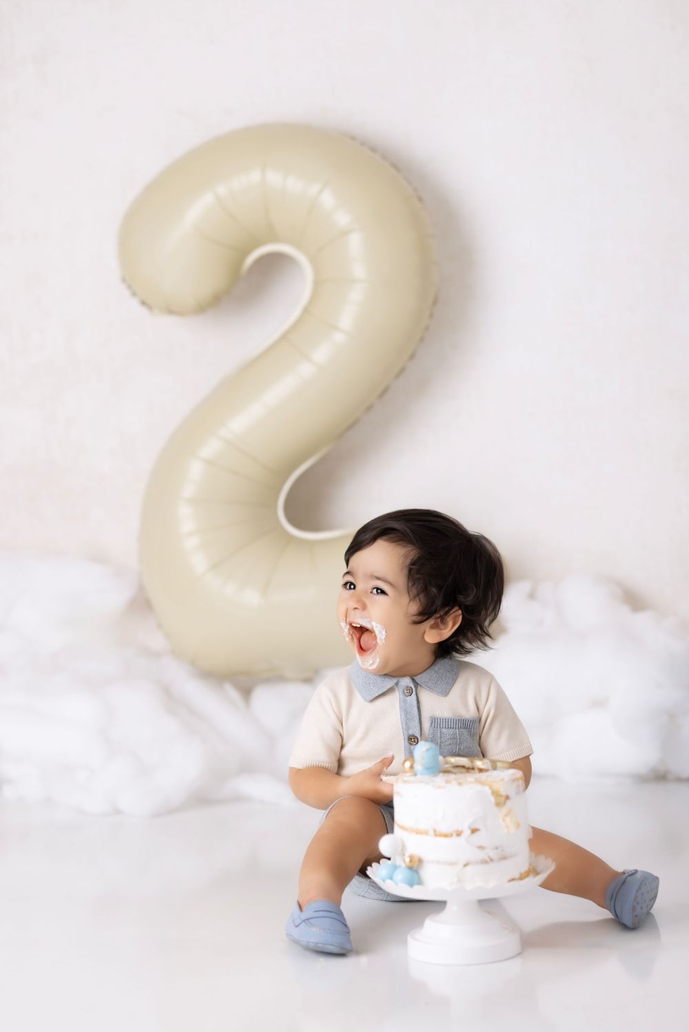A toddler in a cream outfit sits by a large number 2 balloon and small cake during a birthday photoshoot.