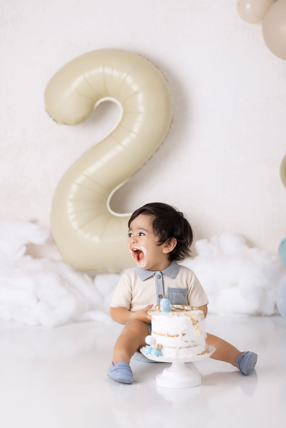 A toddler sits by a large number 2 balloon with a white birthday cake on a pedestal for their second birthday celebration.
