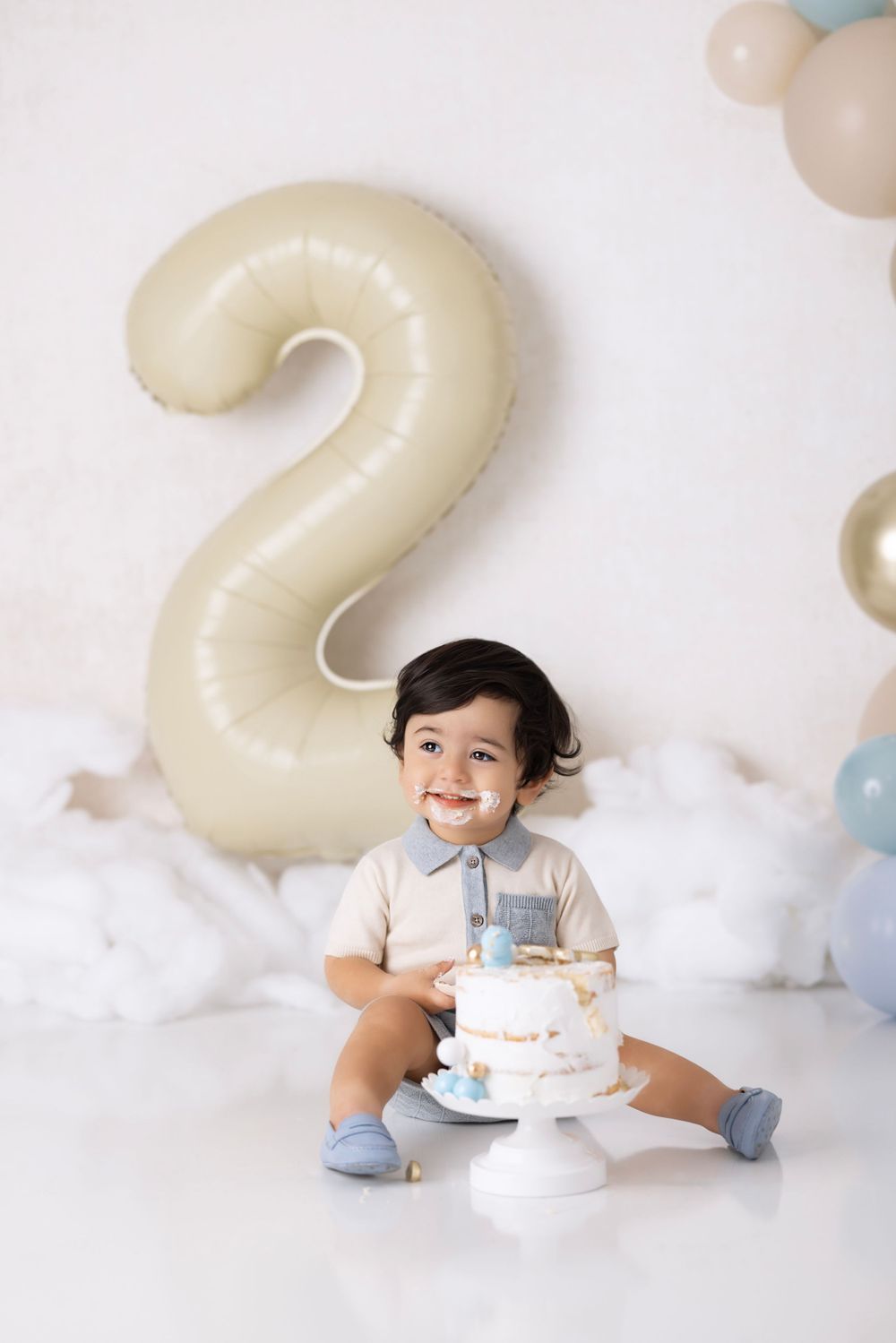 A toddler in a white shirt and blue socks sits with a birthday cake next to large number 2 balloon in a dreamy cloud setting.