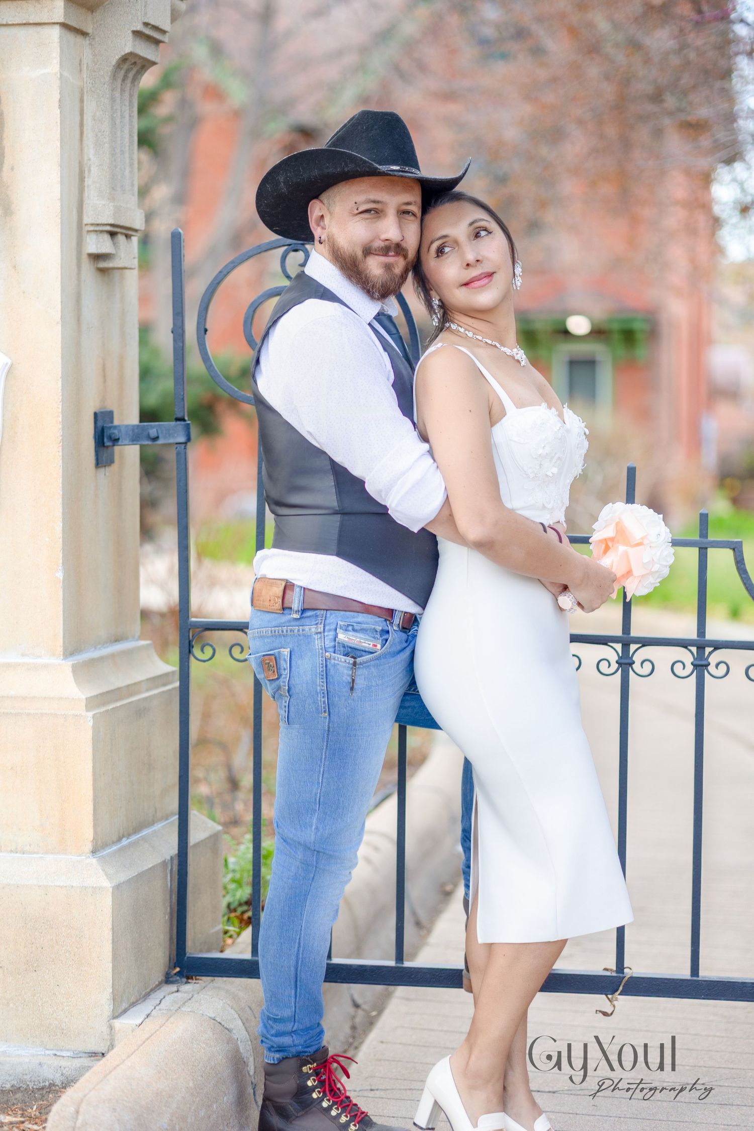 A couple in western-inspired wedding attire pose together against a decorative iron gate and stone pillars outdoors.