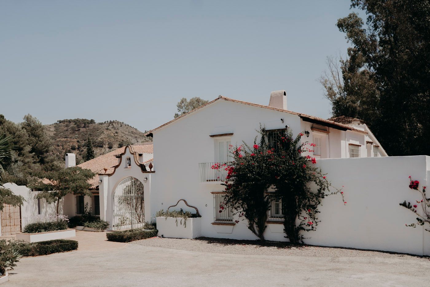 White stucco Spanish-style house with pink bougainvillea flowers and ornate stone archway entrance in a sunny garden setting.