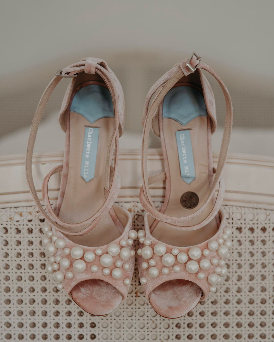 A pair of blush pink peep-toe bridal shoes with pearl embellishments on a woven rattan surface.