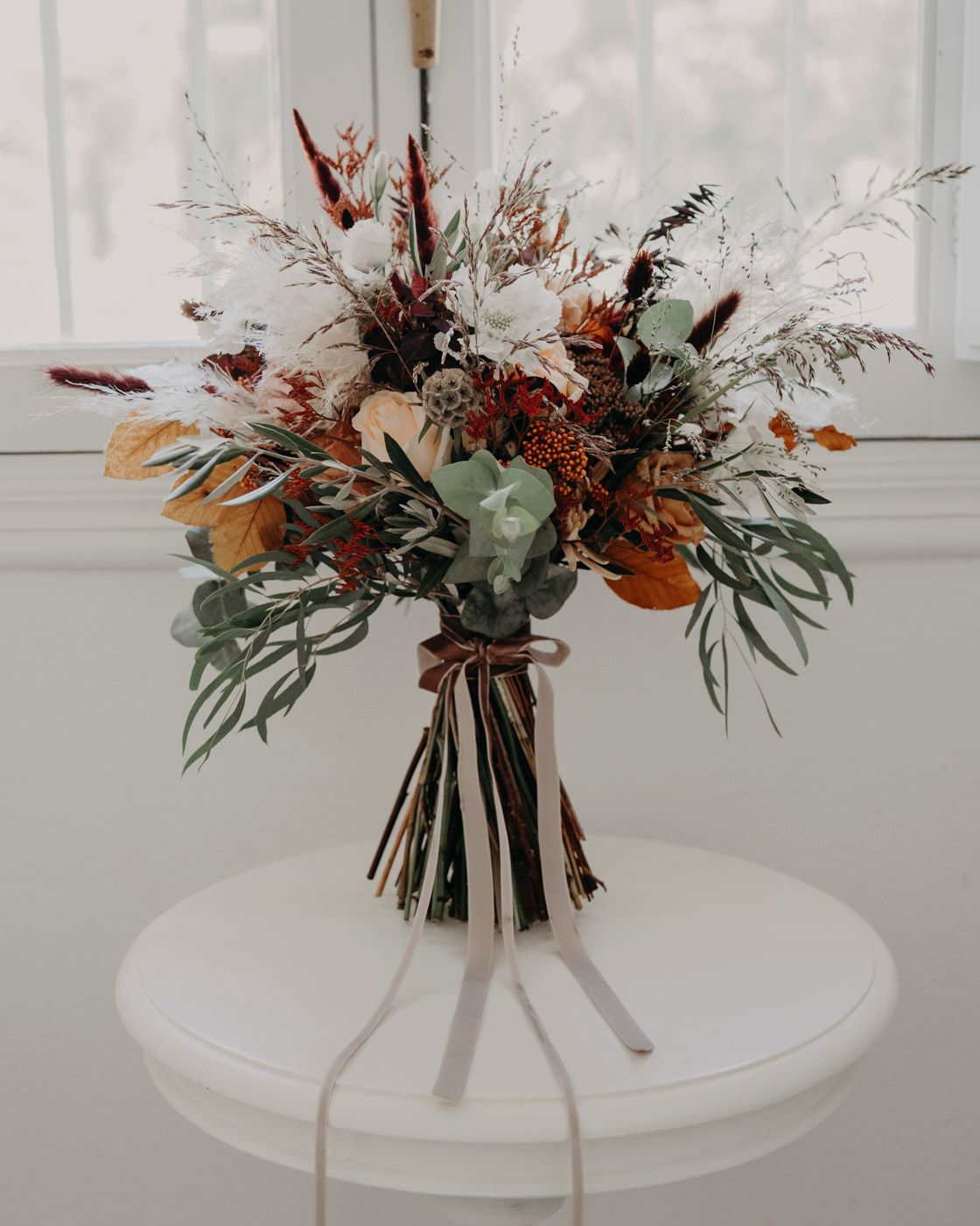 A rustic dried floral bouquet with eucalyptus, burgundy flowers and autumn foliage tied with ribbon on a white surface.
