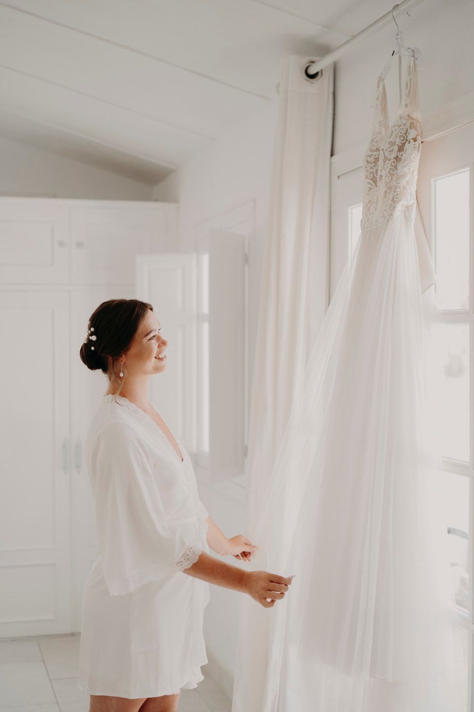 A white wedding dress hangs against a white wall while someone in a robe looks at it in anticipation.