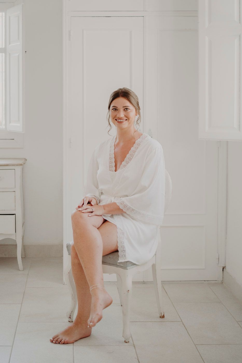 Someone wearing a white robe sits casually on a white chair in a bright minimal room with white tile floors.