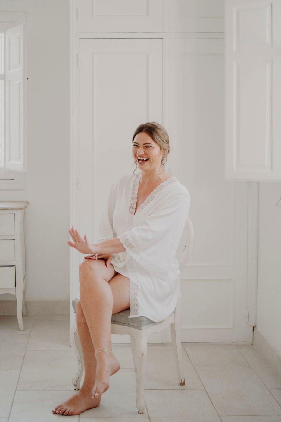 A joyful woman in a white robe sitting on a white chair in a bright minimal room with white walls and floor.