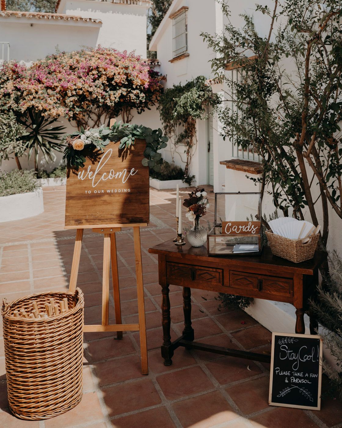 Rustic welcome sign and antique table setup with wicker basket and pink floral decorations in Mediterranean courtyard setting.