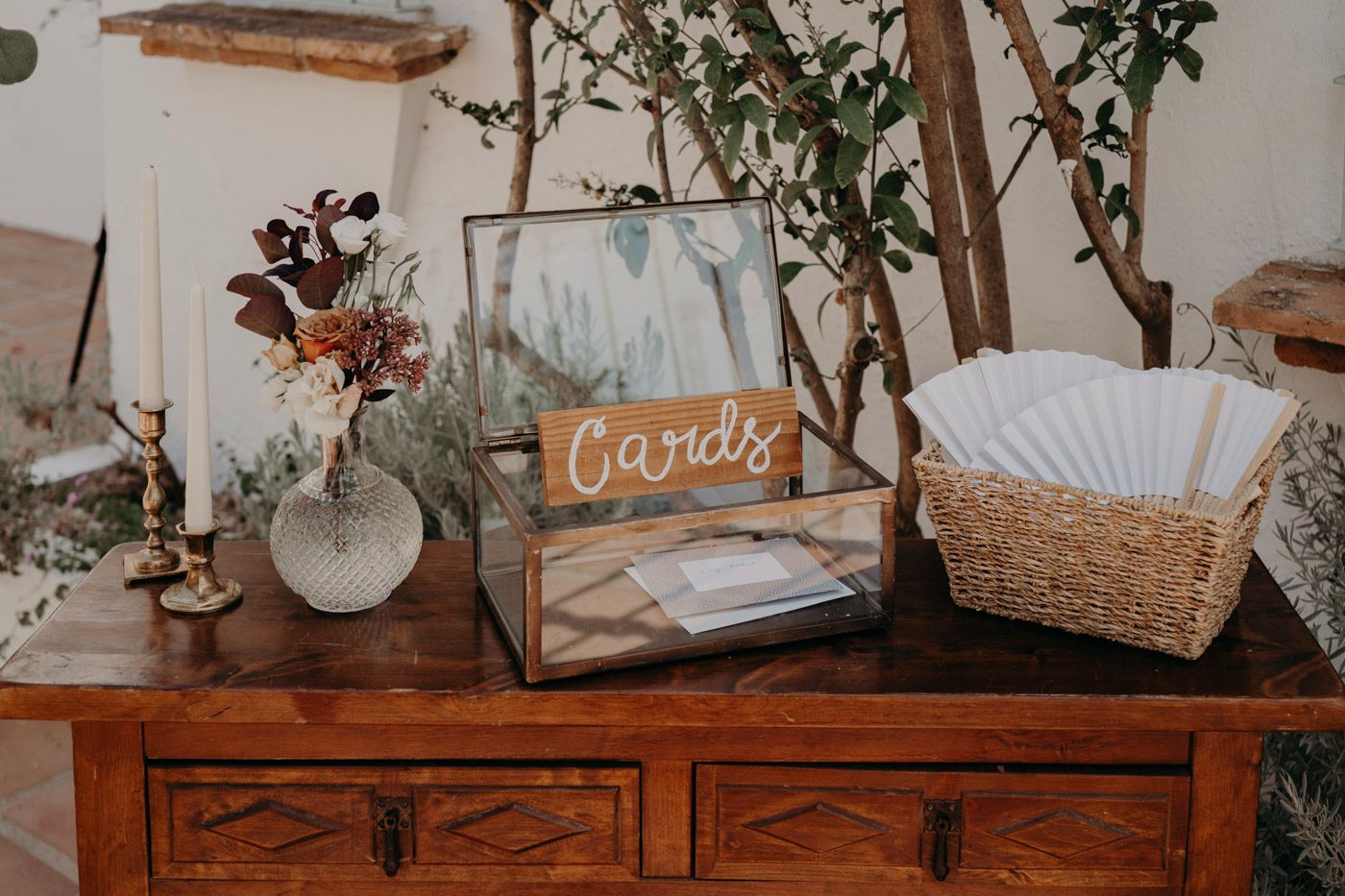 A rustic wedding table display with a wooden 'Cards' sign, glass box, and decorative items on a vintage wooden cabinet.
