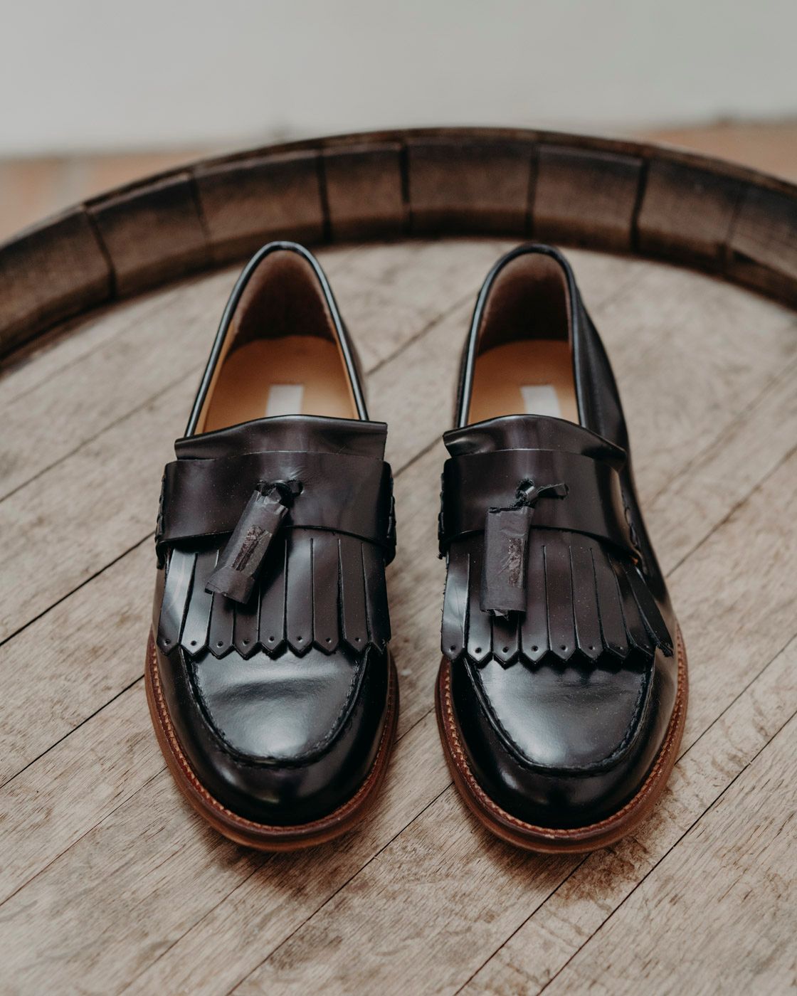 A pair of black leather penny loafers with tassels and fringe displayed on a wooden barrel top surface.