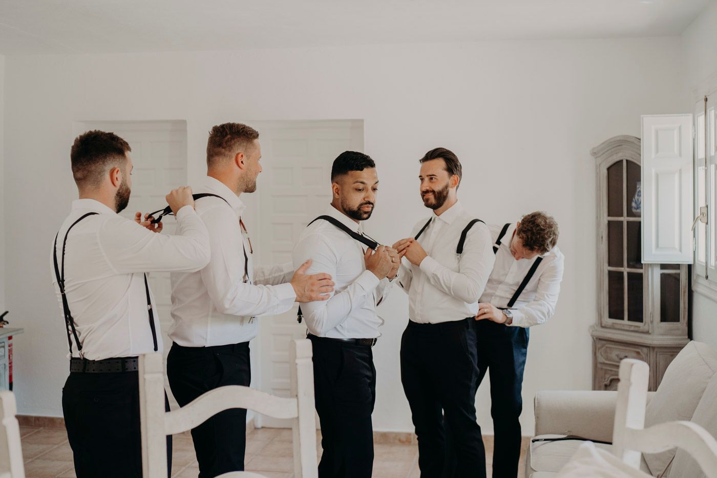 Groomsmen in white shirts and black suspenders share a laugh while getting ready in a bright room.
