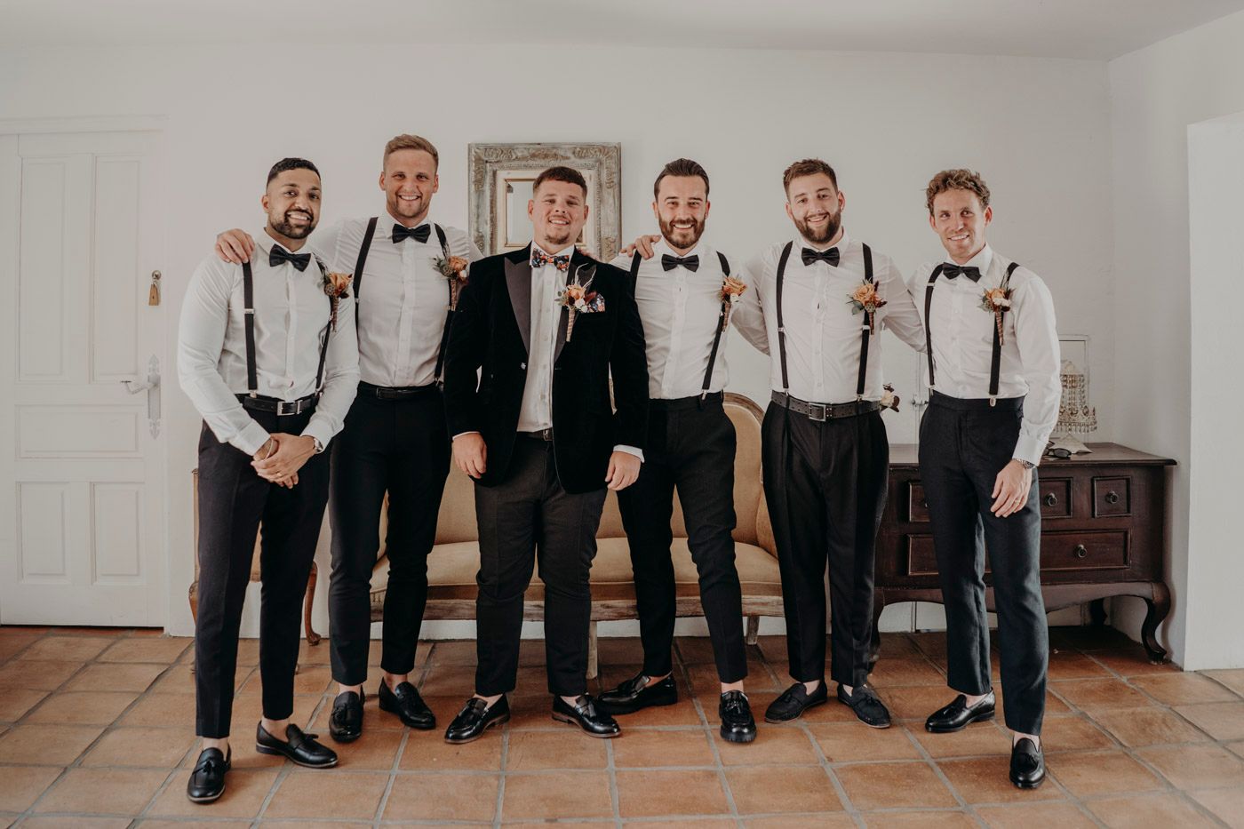 A wedding party of groomsmen wearing black pants with suspenders and bow ties pose together in a light room.