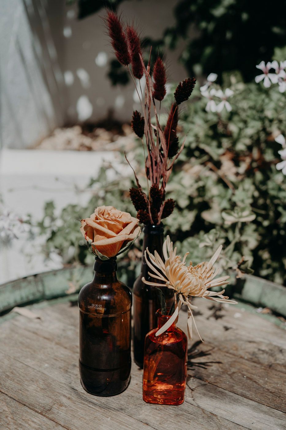 Amber glass bottles with dried flowers and cattails create a moody vintage arrangement on rustic wooden surface.