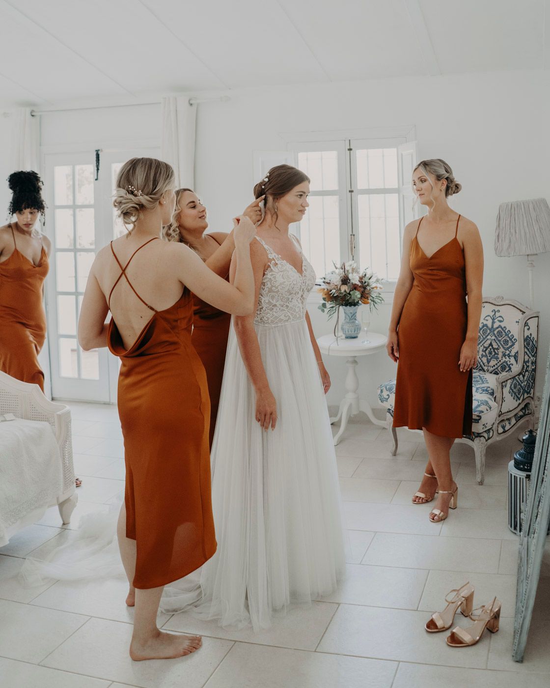 Wedding party getting ready in bright white room with bridesmaids in rust-colored silk dresses helping bride in flowy gown.