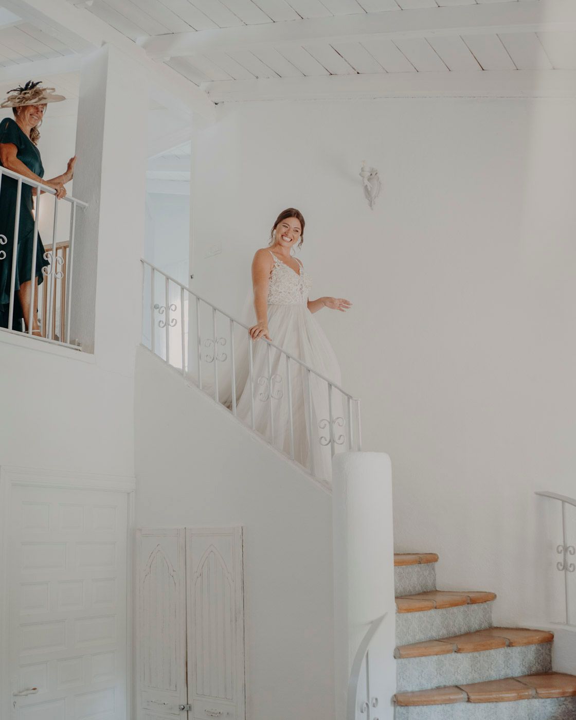 A bride in a white wedding gown stands on a bright staircase while someone watches from above.
