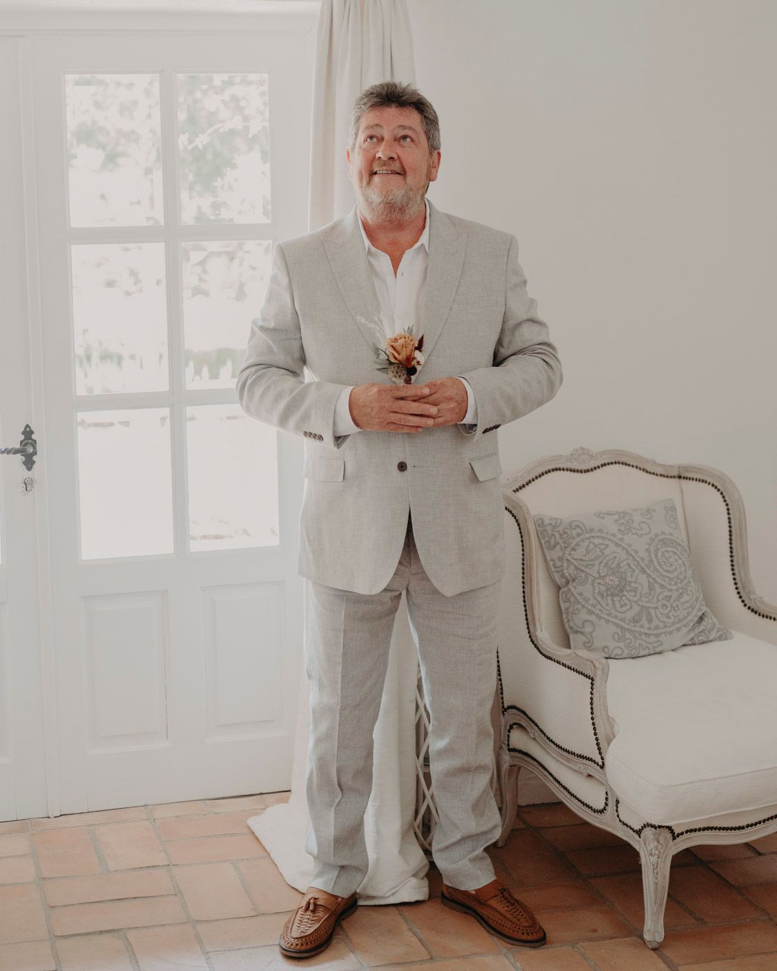 Someone wearing a light gray suit stands in a bright room next to an elegant vintage chair.