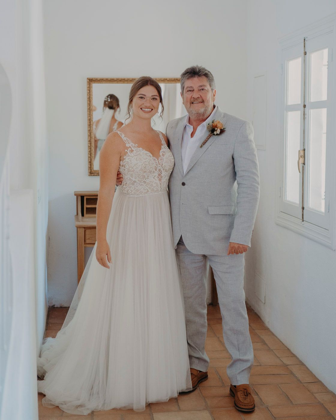 Wedding photo of a couple standing indoors, with the bride in a white gown and groom wearing a light gray suit.