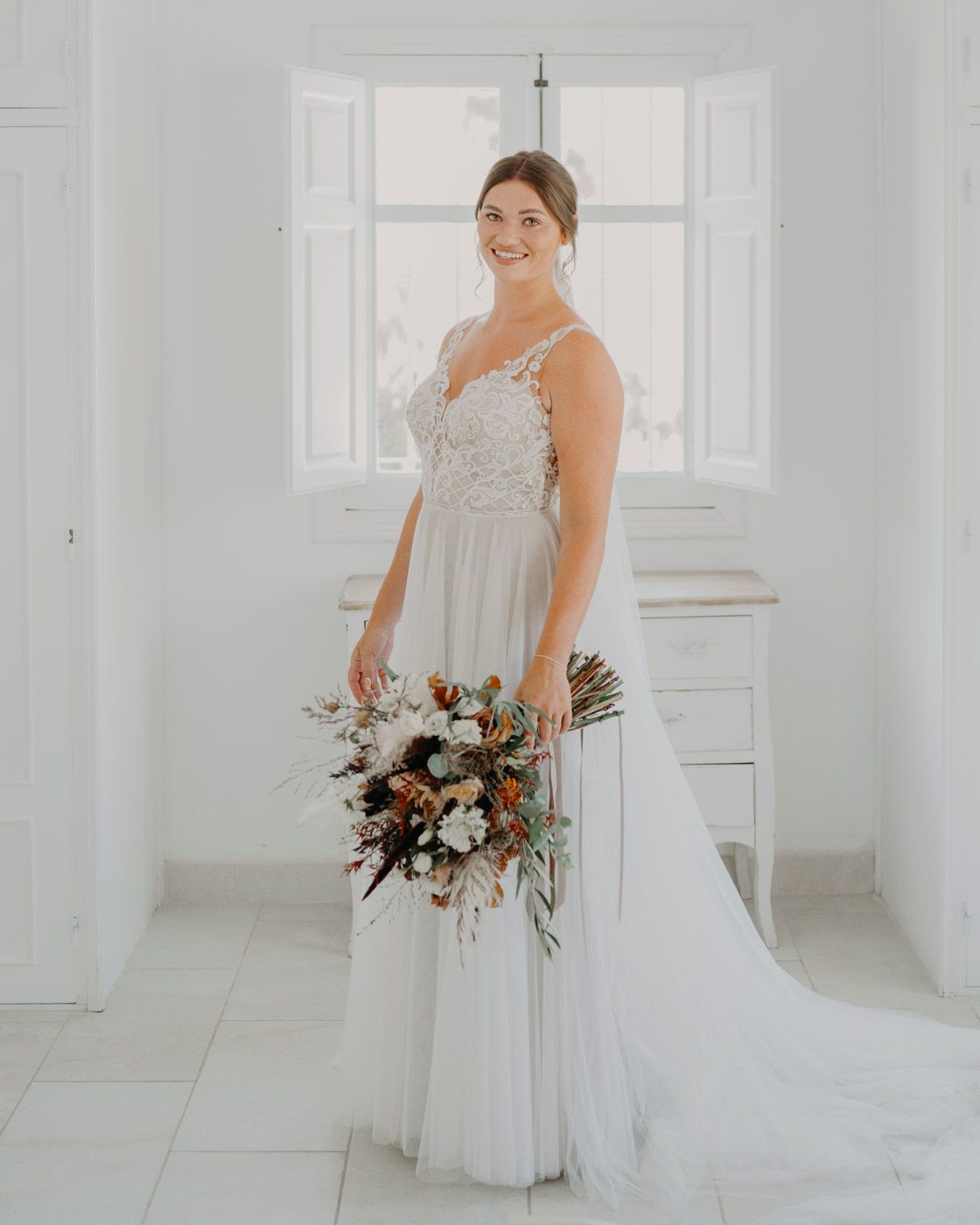 A bride in a white lace wedding gown with a flowing train poses in a bright room while holding a romantic floral bouquet.