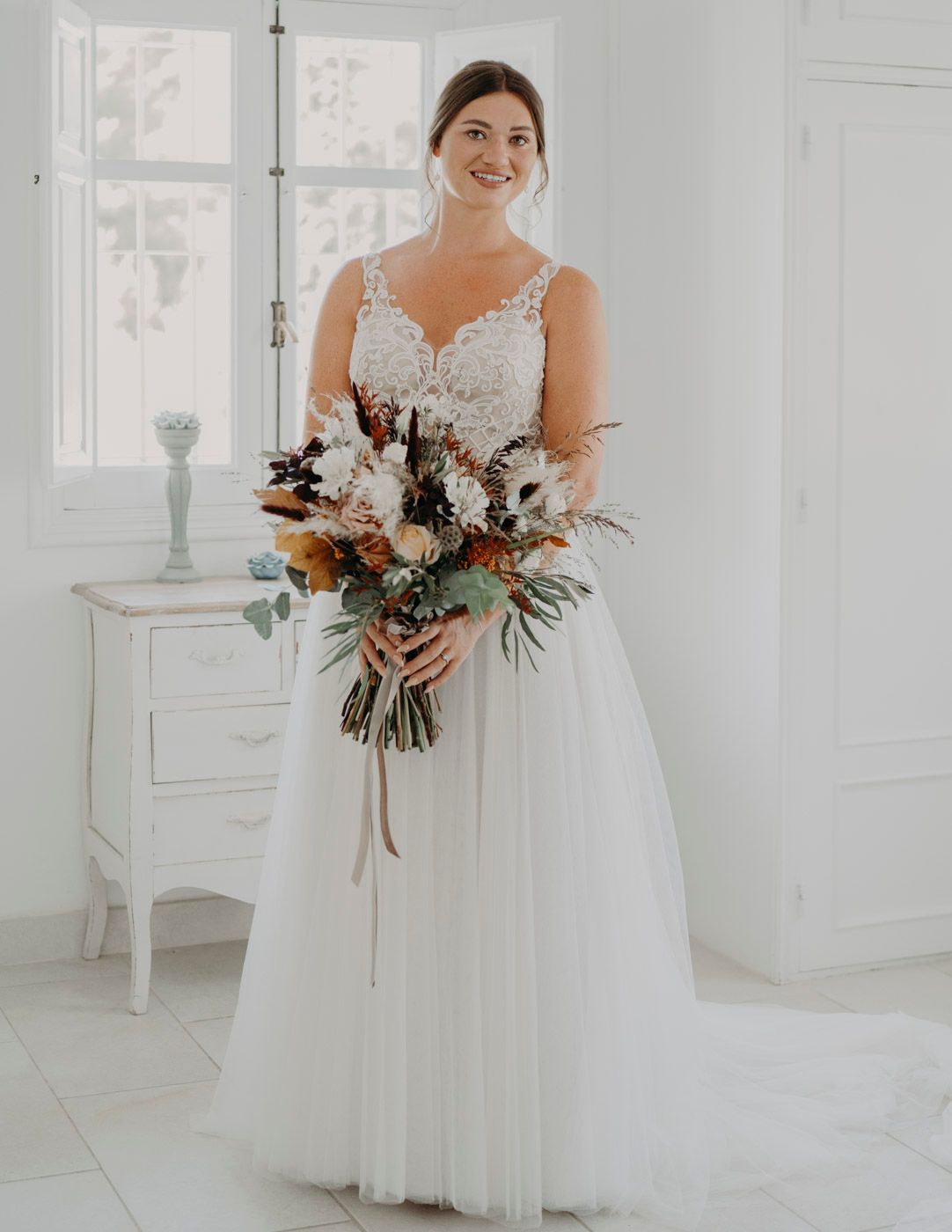 Beautiful wedding dress with lace detail and tulle skirt complemented by rustic autumn-toned floral bouquet in bright room.
