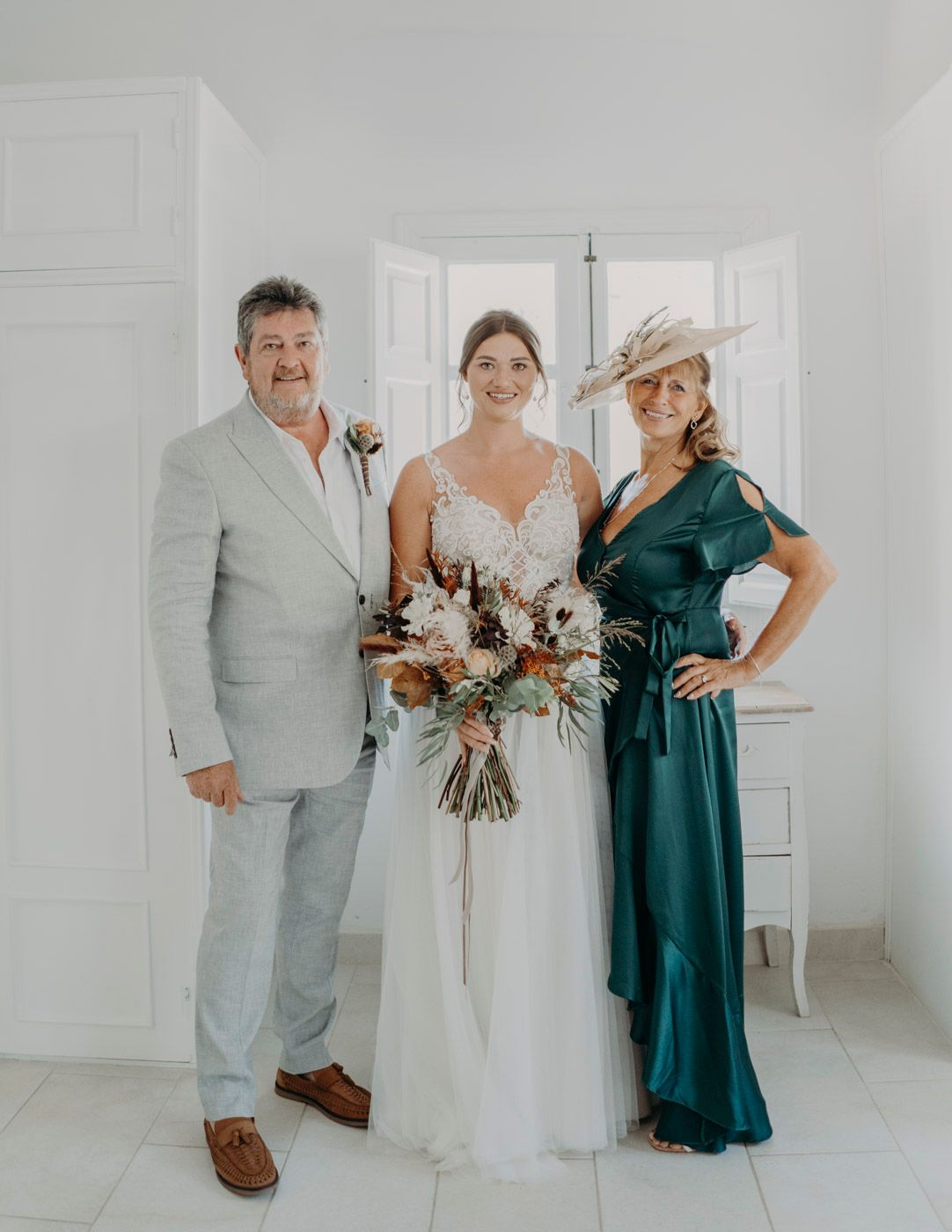 Elegant wedding party photo in white room featuring a bride in lace gown and two people in formal attire.