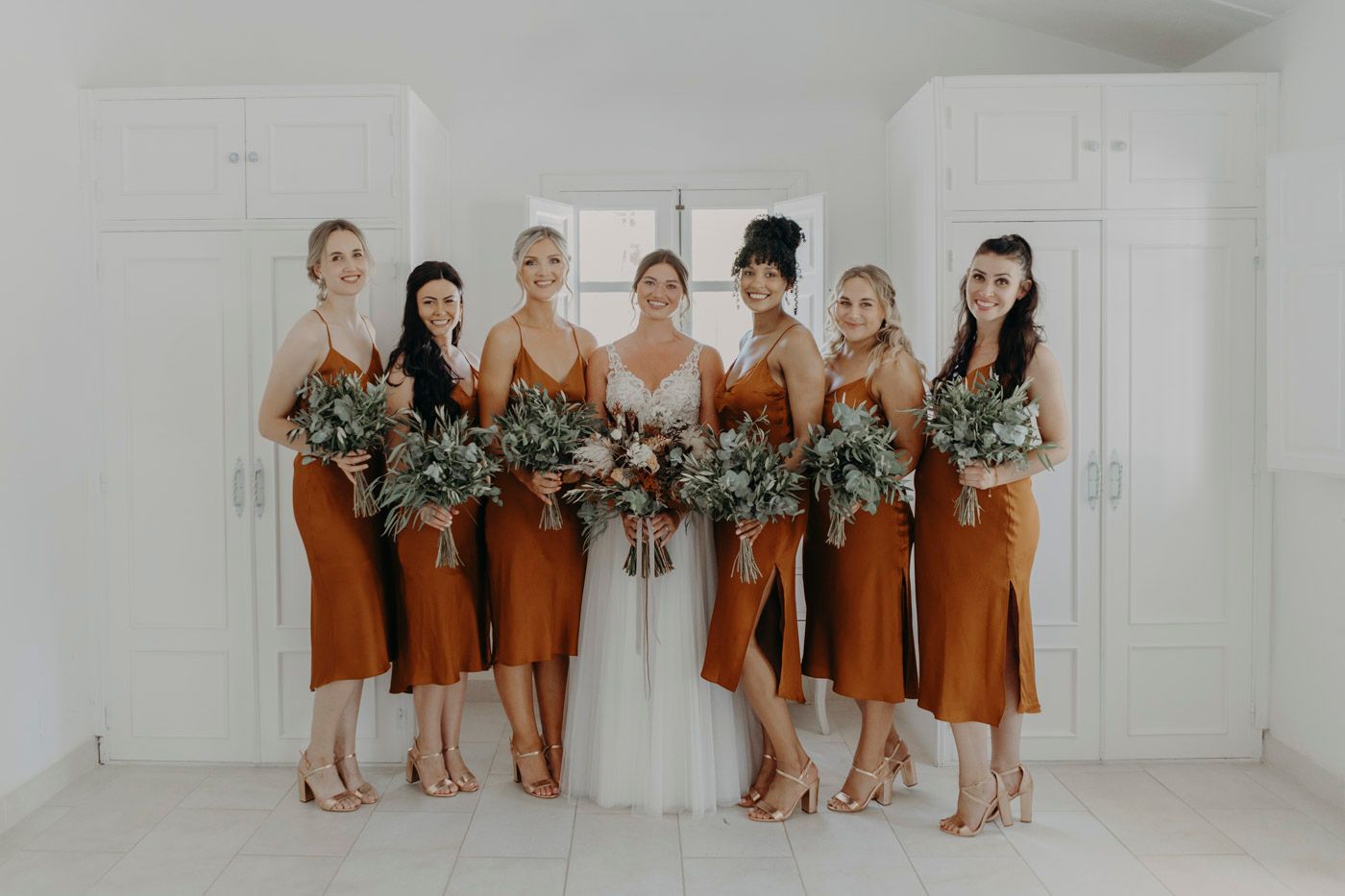 A bridal party wearing rust-colored bridesmaid dresses poses together in a bright white studio with floral bouquets.