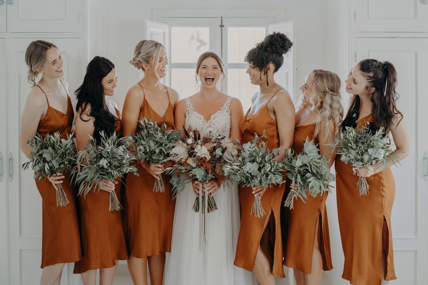Bride in white dress surrounded by bridesmaids wearing matching rust-colored dresses holding greenery bouquets.