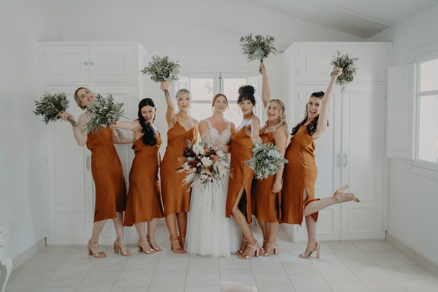 Wedding party in rust-colored bridesmaid dresses and white gown pose playfully in bright white room with floral bouquets.